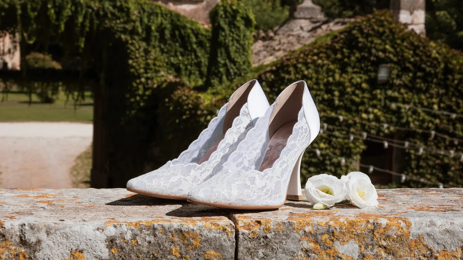Décolleté da sposa Diamante in pizzo bianco appoggiate su un muretto di pietra, con il verde e le rose sullo sfondo