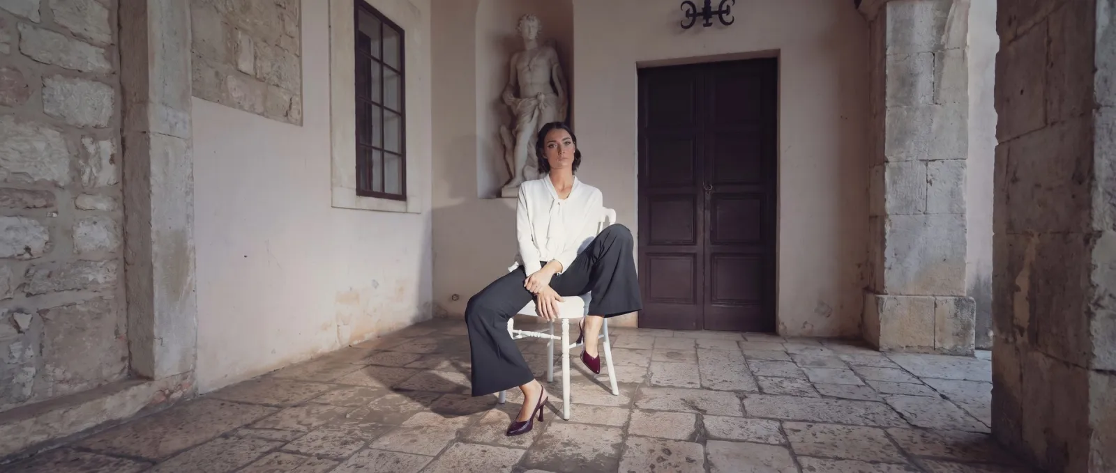 Donna seduta in un cortile in pietra con le slingback Etherea Bordeaux in vernice, fatte a mano a Trani