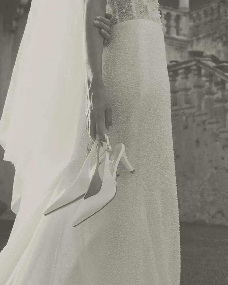Le mani di una sposa tengono due slingback in raso accanto all'abito di paillettes: foto in bianco e nero