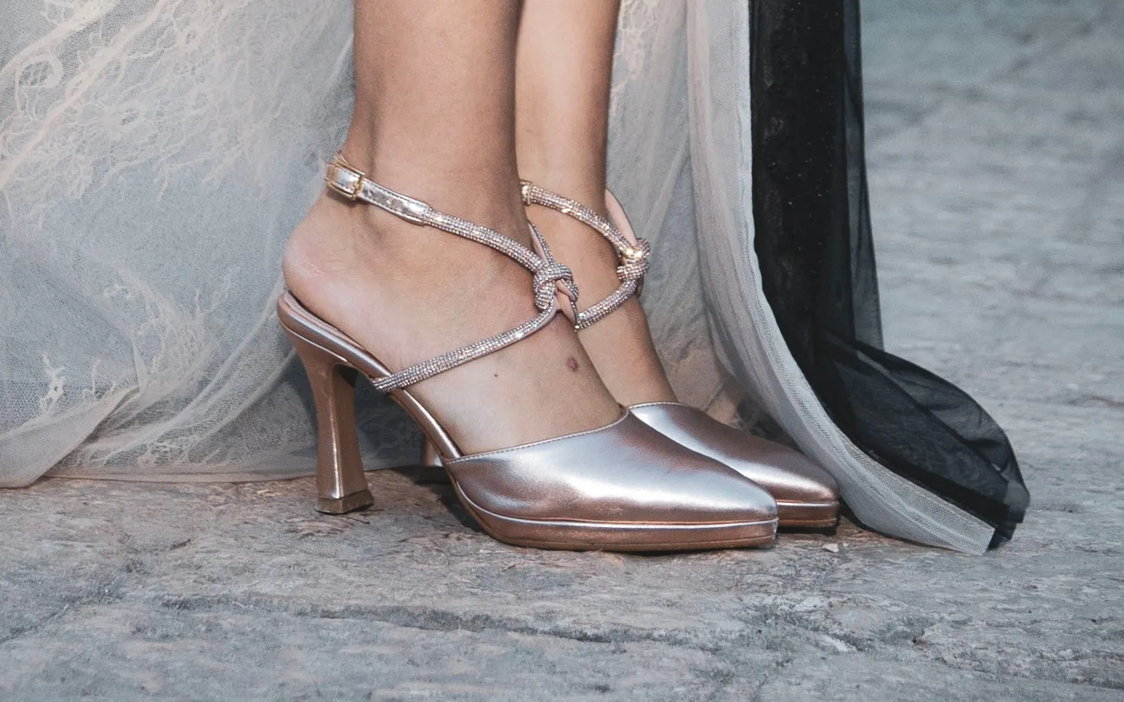 Scarpe rosa metallizzate con il cinturino di strass annodato alla caviglia, accanto all'orlo di un abito in pizzo e tulle