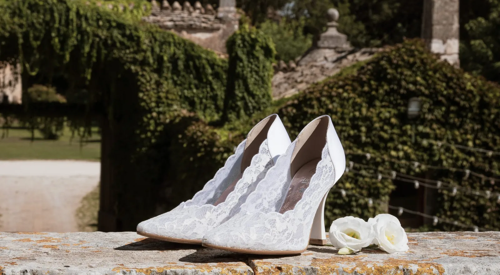 Scarpe da sposa in pizzo appoggiate sul muretto di pietra di una masseria pugliese, con il giardino sullo sfondo