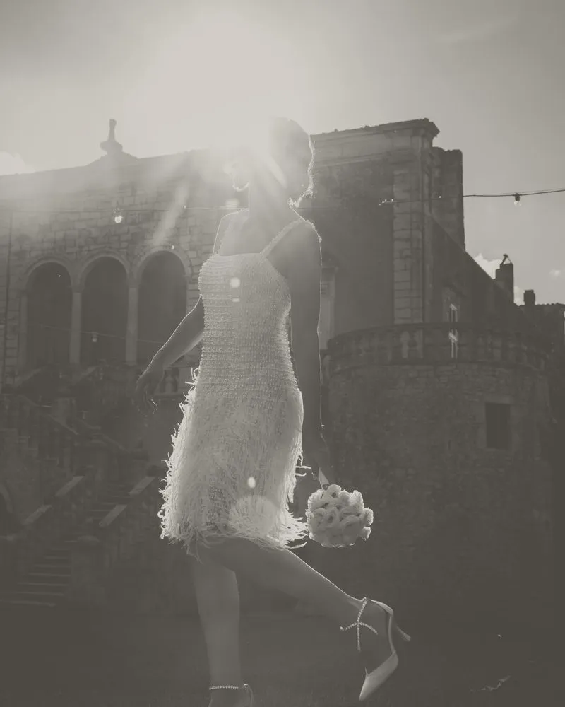 Una sposa in abito corto cammina in controluce davanti a un castello: foto in bianco e nero