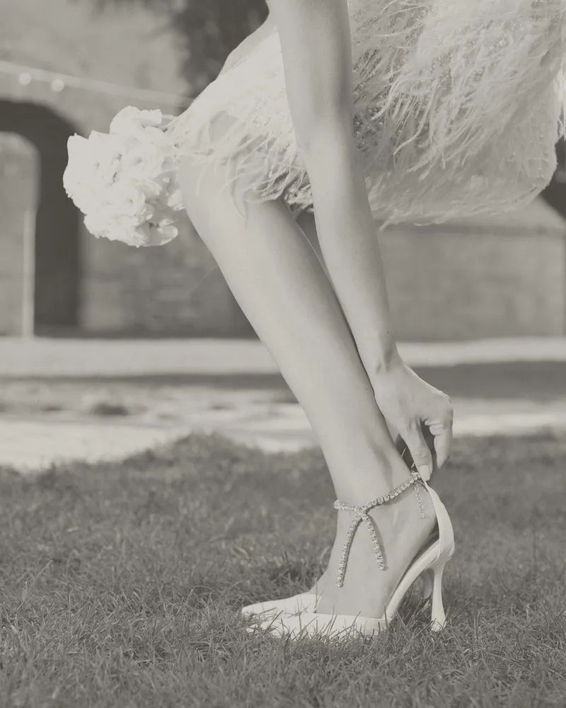 Una sposa allaccia la catenella di un sandalo seduta sul prato: foto in bianco e nero